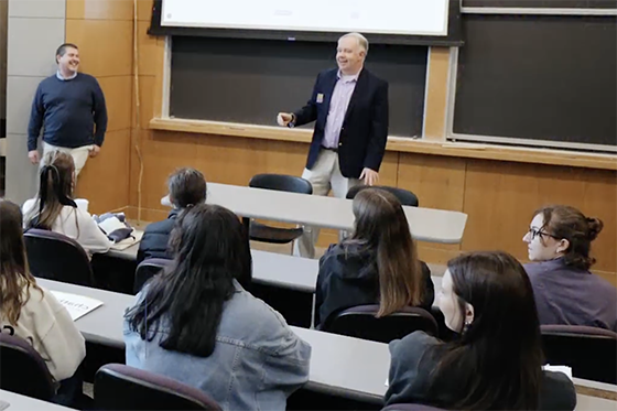 A professor speaks to a group of students seated in a lecture hall, while another professor stands nearby smiling